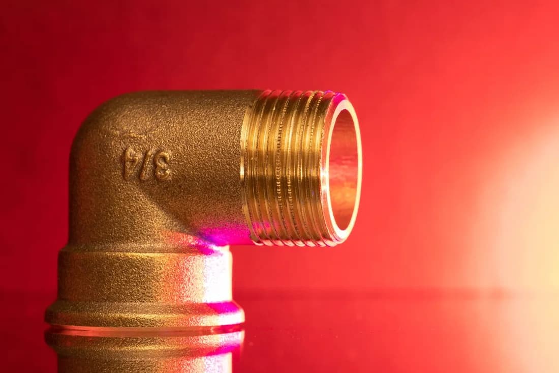 Detailed photograph of a threaded brass elbow pipe fitting against a red background